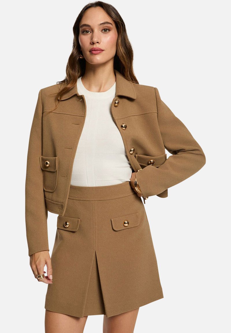 Chaqueta de mezcla de lana color marrón con botones dorados y grandes bolsillos, combinada con una falda marrón a juego que cuenta con un pliegue frontal y bolsillos.