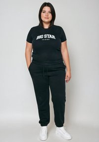 Vrouw staat met handen in zakken, draagt een zwart Big Star Jeans T-shirt, zwarte joggers en witte sneakers tegen een grijze achtergrond.