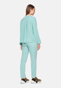Licht turquoise blouse met lange mouwen en een geplooide achterkant, gecombineerd met bijpassende tapered broek en chunky, gemusterde sportschoenen.