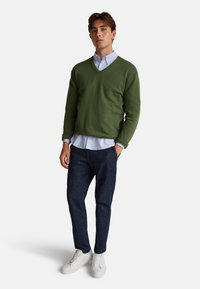 Młody mężczyzna stoi, ubrany w zielony sweter w serek, jasnoniebieską koszulę z guzikami, ciemne dżinsy i białe trampki na białym tle.