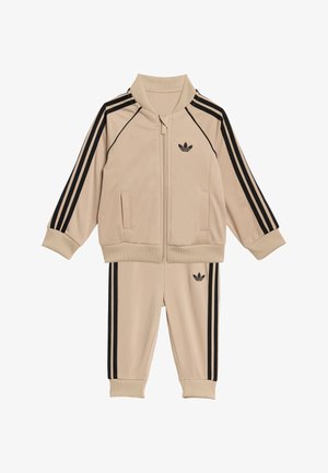 Beige træningsdragt bestående af en lynlåstrøje med sorte striber og matchende bukser, med en lille sort logo på brystet og låret.