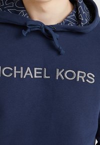 Felpa blu navy con cappuccio e coulisse, foderata all'interno del cappuccio con un motivo a contrasto e logo "MICHAEL KORS" ricamato in argento sulla parte frontale.