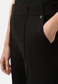 Schwarze Hose mit einer Fronttasche, glatter Textur und vertikalen Nahtdetails. Ein kleines Metall-Logo-Akzent ist am Bund sichtbar.