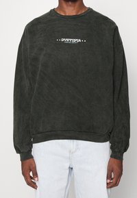 Ciemnozielona bluza z miękkiej tkaniny, z okrągłym dekoltem, ściągaczami na rękawach oraz wyraźnym białym logo na piersi.