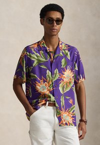 Mężczyzna w okularach przeciwsłonecznych, fioletowej koszuli z krótkim rękawem z dużym pomarańczowo-zielonym wzorem kwiatowym, białych spodniach, brązowym pasku oraz zegarku na nadgarstku.