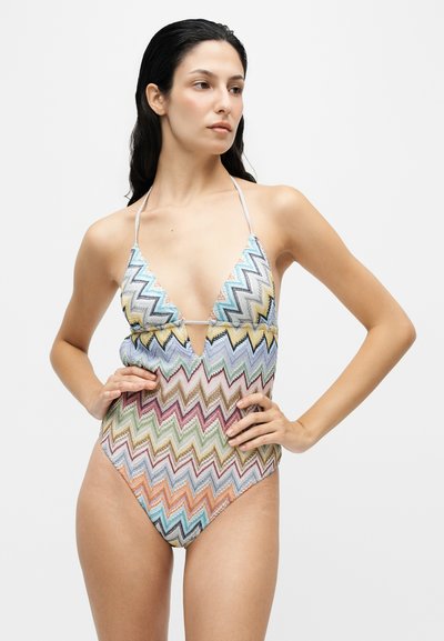 Femme aux longs cheveux foncés porte un maillot de bain une pièce à motif zigzag multicolore avec fines bretelles halter, debout les mains sur les hanches.
