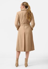 Beige lange jas met een ceintuur in de taille, op maat gemaakt ontwerp en met reverskraag. Glad textuur en knielang, gecombineerd met beige haklaarzen.