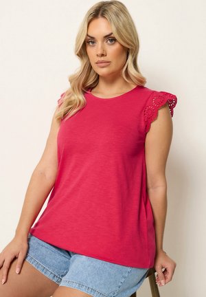 Blondynka w jasnoróżowym bezrękawniku z koronkową lamówką i jasnoniebieskich szortach dżinsowych, siedząca na jednolitym tle.