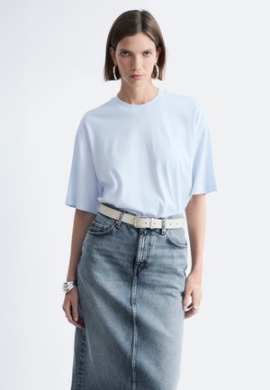 Vrouw met kort bruin haar draagt een lichtblauw oversized T-shirt, creoolringen, zilveren armbanden en een hooggesneden vervaagde denimrok met witte riem.