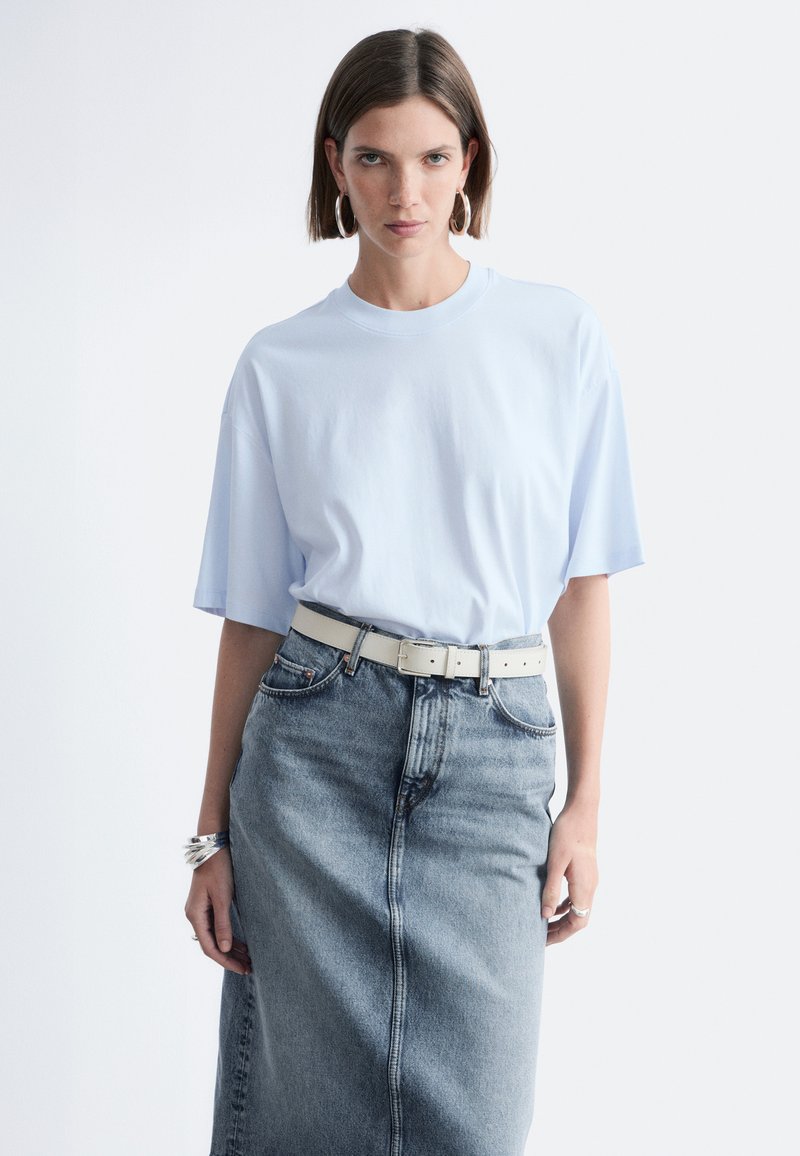 Femme aux cheveux bruns courts portant un T-shirt bleu clair surdimensionné, des boucles d'oreilles créoles, des bracelets en argent et une jupe en jean délavée taille haute avec une ceinture blanche.