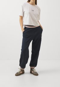 Bílý zkrácený tričko s cvočkovým designem a logem EA7; tmavě modré tepláky s elastickým pásem; vzorované tenisky se šněrováním.