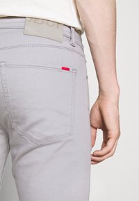 Pantalones de mezclilla de color gris claro, que cuentan con un bolsillo trasero con una pequeña etiqueta roja y un parche de cuero gris claro en la cintura.
