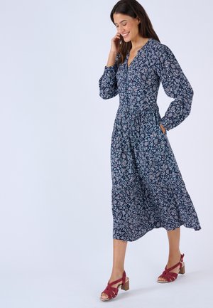 Mujer sonriente caminando con un vestido midi azul marino con estampado floral, mangas largas y sandalias rojas de tacón grueso, sobre un fondo liso.