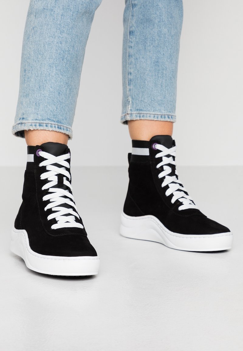 Baskets montantes en daim noir avec des lacets blancs et une semelle plateforme blanche. Elles présentent une sangle noire avec un accent gris à la cheville.