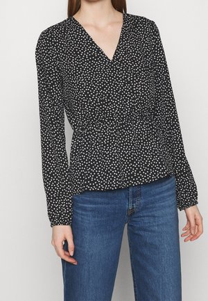 Vrouw draagt een zwarte blouse met lange mouwen met witte hartjesprint en blauwe spijkerbroek, staand tegen een effen lichte achtergrond.