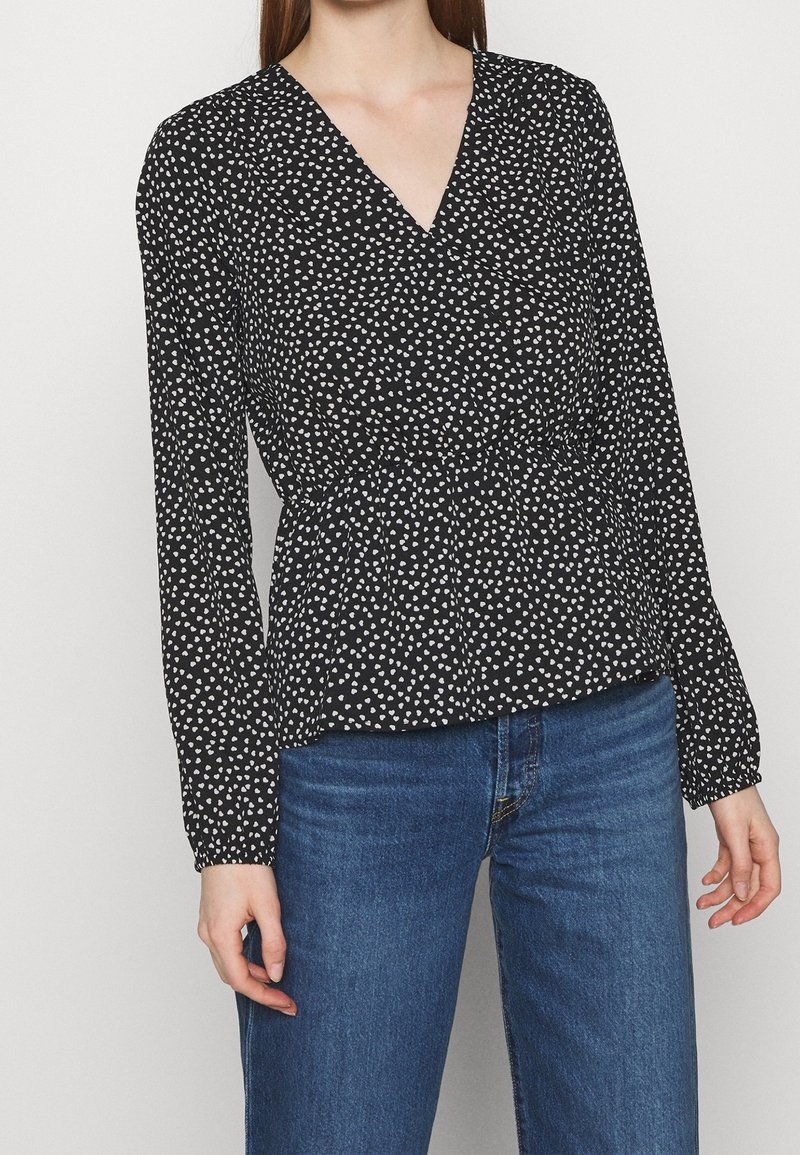 Femme portant un chemisier noir à manches longues avec un imprimé cœur blanc et un jean bleu, debout devant un fond clair uni.