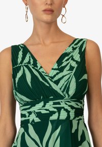 Robe verte avec un corsage plissé et un décolleté en V, ornée de motifs légers de feuilles vertes, taille cintrée et jupe fluide. Boucles d'oreilles en or en forme de cercle.