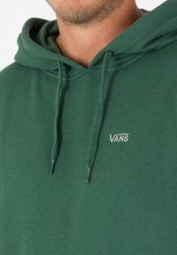 Zelená mikina s kapucí z měkké bavlny. Má klokaní kapsu, stahovací šňůrku na kapuci a malý vyšívaný logo Vans na hrudi.