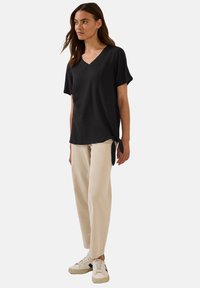 Blusa nera a scollo a V con maniche corte, caratterizzata da un dettaglio con nodo laterale. Abbinata a pantaloni beige chiaro e sneakers bianche.