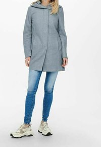 Abrigo gris con capucha, corte recto y bolsillos laterales, combinado con vaqueros azules y zapatillas de color claro. Textura de tela lisa, diseño minimalista.