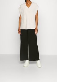 Helles beige gestricktes Sweatshirt mit V-Ausschnitt und kurzen Ärmeln, kombiniert mit weit geschnittenen schwarzen Hosen und weißen Sneakers.