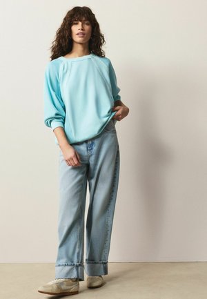 Frau mit lockigem Haar, die einen hellblauen oversized Pullover, lockere hellblaue Jeans mit aufgerollten Bündchen und cremefarbene Sneakers trägt, steht drinnen.