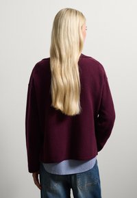 Bordeaux trui met een ontspannen pasvorm, ronde halslijn en geribbelde onderkant, gedragen over een lichtblauw geruit shirt, gecombineerd met vervaagde blauwe jeans.