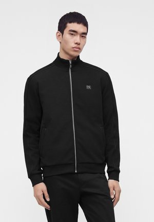 Jeune homme portant une veste noire zippée avec col montant et un pantalon noir assorti, debout devant un fond clair uni.
