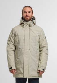 Beige isolerad parka med hög krage, justerbar huva och sidofickor. Har en mjuk textur och enkel design.