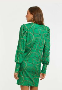 Vihreä mekko, jossa on pitkät puffihihat ja istuva siluetti. Siinä on kultainen ja musta ketjumalli sileällä kankaalla.