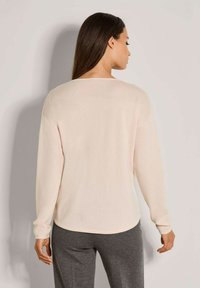 Frau mit langen dunklen Haaren trägt einen hellbeigen langärmligen Pullover und eine dunkelgraue Hose, von hinten gegen einen einfarbigen Hintergrund gezeigt.