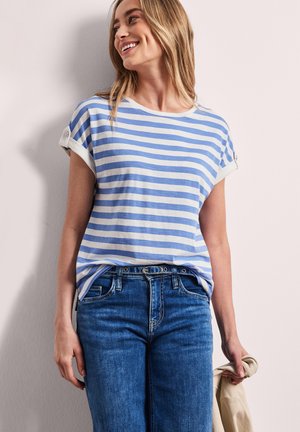 Femme souriante, portant un t-shirt à manches courtes rayé bleu et blanc et un jean bleu, tenant une veste beige dans sa main droite.