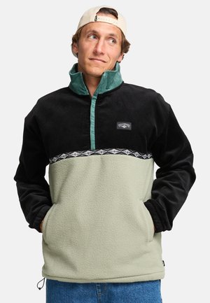 Fleece pullover in black, green, and light beige. Features a high collar, horizontal stripe design, and a branded patch. Textured material.