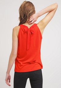 Top rosso senza maniche realizzato in un tessuto morbido, con colletto alto e dettaglio a fiocco sulla schiena. Design semplice e aderente, senza motivi visibili.