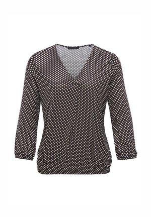 Kobieta ubrana w czarno-białą bluzkę w wzory, jasnoniebieskie skrócone dżinsy z surowymi brzegami oraz białe tenisówki na sznurówki, ręce w kieszeniach.