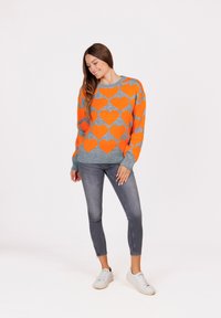 Grauer Pullover mit lebhaften orangefarbenen Herzmustern, rundem Ausschnitt und gerippten Bündchen, kombiniert mit grauen Slim-Jeans und weißen Sneakers.