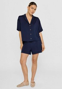 Marineblaues Kurzarmhemd mit Knöpfen und passende Shorts. Weicher Stoff, lockerer Schnitt, Seitentaschen und einfaches Design. Tragen mit beigen Slippern.