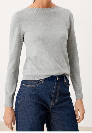 Kvinde iført en lysegrå strikket trøje med lange ærmer og mørkeblå højtaljede jeans, stående med hænderne afslappet ned langs siderne.
