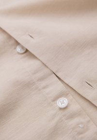 Gros plan d'un tissu de chemise beige montrant des boutons blancs et des boutonnières le long de la patte de boutonnage avant.