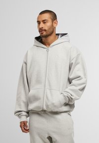 Hombre con barba y cabello corto, vistiendo una sudadera con cremallera color gris claro y pantalones de chándal a juego, de pie con una mano en el bolsillo.