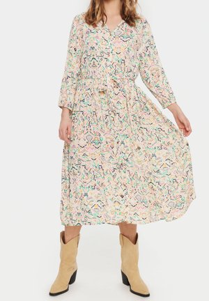 Nainen pukeutunut pitkähihainen, pastellikuosinen midimekko, jossa on vyötärösolki, yhdistetty beigeihin cowboy-saappaisiin, seisoo yksiväristä taustaa vasten.