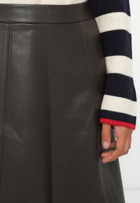 Jupe en cuir marron avec une texture lisse et un design évasé. Une main repose sur la jupe, portant un pull rayé rouge, marine et blanc.
