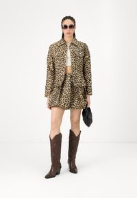 Set s kratkim leopardjim potiskom, ki vključuje jakno in krilo, z bež ozadjem in temnimi pikami. Temno rjavi kavbojčki in črna torbica dopolnjujeta videz.