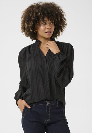 Schwarze Bluse mit vertikalen Texturstreifen, Rüschenspalte und Knopfverschluss. Lange Ärmel mit gerafften Bündchen. Kombiniert mit dunklen Jeans.
