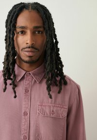 Langärmliges Shirt aus mauvefarbenem Stoff, mit klassischem Kragen, Knöpfen auf der Vorderseite und einer Brusttasche. Glatte Textur mit taillierter Passform.