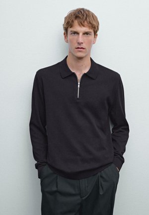 Young man with light brown hair wearing a black long-sleeve zippered polo shirt and dark trousers, standing against a light gray wall.