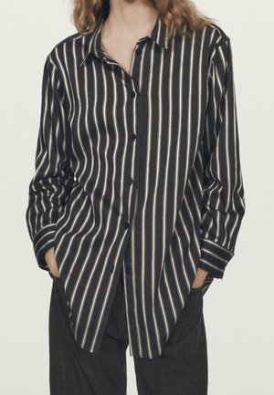 Personne portant une chemise noire boutonnée avec des rayures blanches verticales et un pantalon noir, mains décontractées dans les poches, debout devant un fond uni.