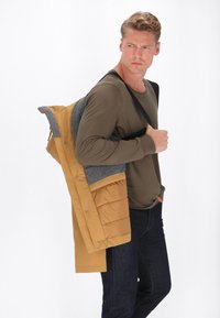 Geltonas pūkinis džemperis su pilkais akcentais, turintis tekstūrinį paviršių ir spaustukus. Dėvimas ant ilgo rankovės alyvuogių spalvos marškinėlių ir tamsių džinsų.