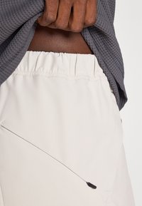 Calções de desporto bege claro com um cós elástico e um detalhe de costura diagonal. Um fecho éclair preto fino é visível no lado direito.