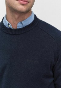 Marinblå stickad tröja med ribbat rundhals. Har subtilt broderad logotyp på bröstet och en slät yta.